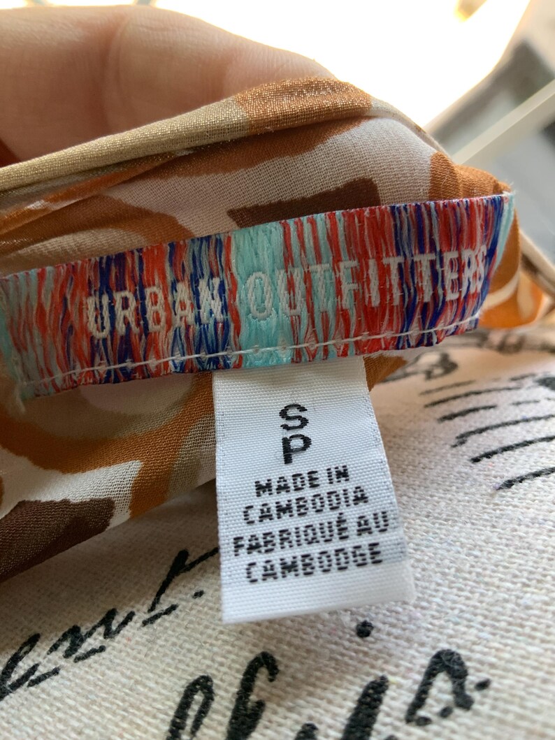 K&ouml;nnte beinhalten: Nahaufnahme eines Etiketts von Urban Outfitters. Das wei&szlig;e Etikett tr&auml;gt den schwarzen Text "S", "Made in Cambodia" und "Fabriqu&eacute; au Cambodge". Der Stoff hat ein braunes und wei&szlig;es Muster.