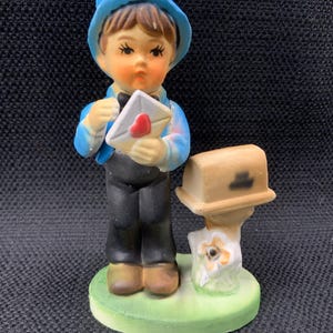 Puede incluir: Una figurita de un niño con un sombrero azul y una camisa azul que sostiene una carta con un corazón rojo. El niño está de pie junto a un buzón con una flor blanca en la base.