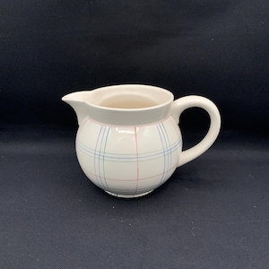 May include: A white ceramic creamer with a round body and a handle. The creamer features a plaid pattern in red, blue, and green lines. The spout is on the left side of the creamer.