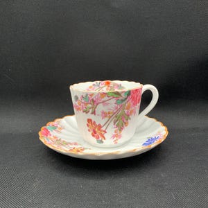 May include: A white porcelain teacup and saucer set featuring a scalloped edge and a delicate floral pattern in shades of pink, orange, green, and blue. The rim of both the cup and the saucer is finished with a thin, metallic gold-colored trim.