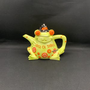 May include: A whimsical, lime green frog-shaped teapot with orange accents and a witch's hat. The teapot features the words "ABRA CA DABRA" and decorative swirls. A unique and fun addition to any kitchen or tea collection.