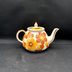 Puede incluir: Una tetera de cerámica blanca con un patrón floral pintado en tonos amarillos, naranjas y marrones. La tetera tiene un asa curva, un pico vertedor cónico y una tapa extraíble rematada con un remate puntiagudo de color dorado.