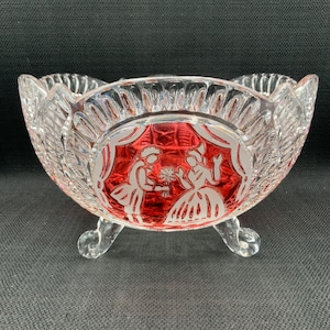 Può includere: Una ciotola di vetro trasparente con una base rossa e un disegno inciso bianco di una coppia. La ciotola ha tre gambe e un bordo smerlato.