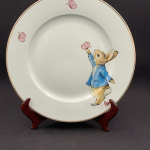 Puede incluir: Un plato de cerámica blanco con un borde dorado presenta una representación de Peter Rabbit con una chaqueta azul, alcanzando una mariposa rosa. Dos mariposas rosas adicionales se representan en el plato.