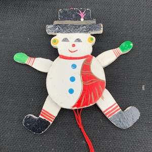 May include: A wooden snowman ornament with a black hat, green mittens, and red and white striped legs. The snowman has a red scarf and a red string attached. The snowman has a smiling face with yellow earrings and blue buttons.