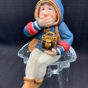 Op de afbeelding: Een blauwe en rode figuur van een kabouter die op een glazen standaard zit. De kabouter houdt een klein houten doosje vast met een gouden slot en het woord "Secrets" erop. De kabouter houdt zijn hand voor zijn mond.