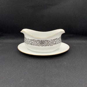 May include: White gravy boat with a black floral design and gold trim, resting on a matching saucer. The gravy boat has a curved handle and a spout for easy pouring. The set is made of porcelain and is suitable for serving sauces and gravies.