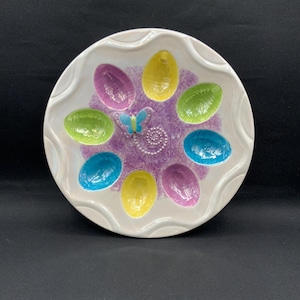 Peut inclure: Un plat de service en céramique blanche pour œufs de Pâques avec un bord festonné. Le plat présente huit empreintes en forme d'œuf colorées dans des tons de bleu, vert, jaune et violet, avec un papillon et un motif tourbillonnant au centre.