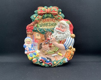 Może przedstawiać: Dekoracyjna ceramiczna tablica ścienna z napisem Santa's Workshop, przedstawiająca rzeźbionego Świętego Mikołaja malującego konika na biegunach. Scena zawiera lalkę, samochodzik, zapakowany prezent oraz skarpetę z napisem Dec 24 w zielonej girlandzie.
