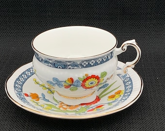 May include: A white teacup and saucer with a floral pattern in shades of blue, red, yellow, and green. The cup has a gold rim and the saucer has a gold band around the edge.