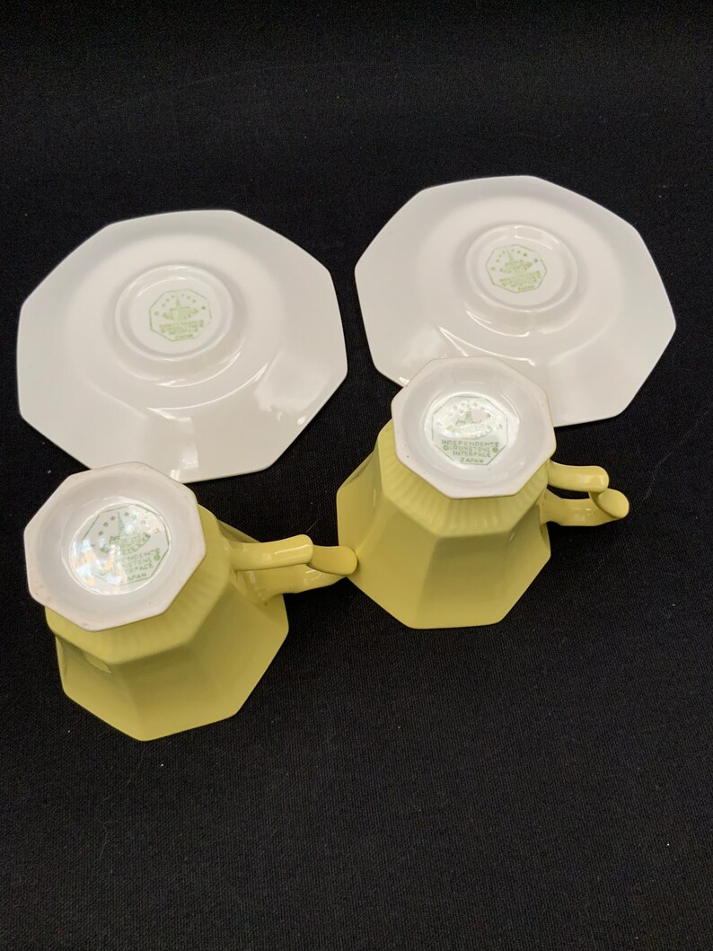K&ouml;nnte beinhalten: Zwei gelbe Teetassen und Untertassen mit einem wei&szlig;en achteckigen Design. Die Tassen und Untertassen haben ein gr&uuml;nes Blumenmuster auf der Unterseite.