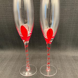 Op de afbeelding: Twee heldere champagneglazen met rode accenten. De stelen zijn versierd met rode spiraalvormige ontwerpen en de basis van elk glas heeft een rood hartvormig ontwerp. De glazen staan tegen een donkere achtergrond.
