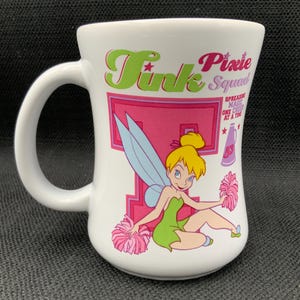 Peut inclure: Une tasse en céramique blanche avec un motif rose, vert et bleu représentant la fée Clochette, le texte "Tink Pixie Squad" et la phrase "Spreading Magic One Pixie At A Time".