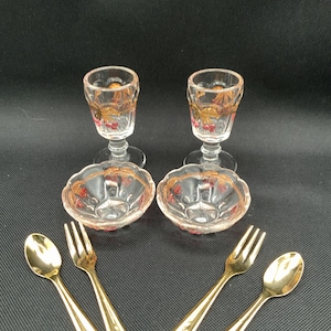 Könnte beinhalten: Zwei klare Glaspokale mit Stiel und zwei passende kleine Glasschalen, jeweils verziert mit goldenen Blattmustern und roten Beerenakzenten. Das Set enthält vier goldfarbene Metallbestecke, bestehend aus zwei Löffeln und zwei Gabeln mit strukturierten Griffen.