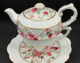 May include: A white porcelain tea set with a teapot, teacup, and saucer. The set is decorated with pink roses and gold trim. The saucer has a scalloped edge. The teapot has a lid and a handle. The teacup sits inside the teapot.