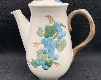 May include: White ceramic teapot with a lid, spout, and handle. The teapot is decorated with a blue and green grape design. The handle and the top of the lid are brown. The teapot is on a black surface.