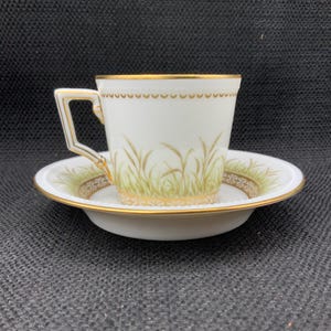 May include: White teacup and saucer set with gold trim and a wheat design. The teacup has a square handle and the saucer has a matching wheat and gold border. The set is likely porcelain or fine china.