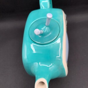 May include: A turquoise ceramic teapot with a handle, spout, and lid. The lid has two light purple knobs. The teapot has a rounded shape with a white and light blue accent on the side. The teapot is designed for brewing tea.