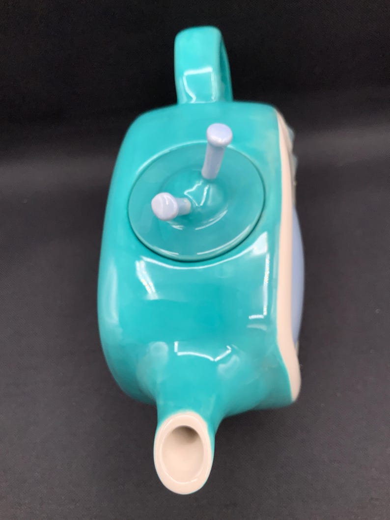 May include: A turquoise ceramic teapot with a handle, spout, and lid. The lid has two light purple knobs. The teapot has a rounded shape with a white and light blue accent on the side. The teapot is designed for brewing tea.