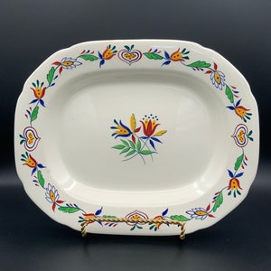 Puede incluir: Una fuente ovalada blanca con un diseño floral en rojo, amarillo, azul y verde. El diseño floral está alrededor del borde de la fuente y presenta flores y hojas estilizadas.