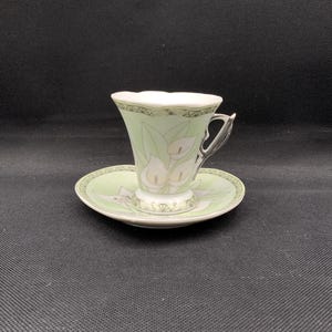 Könnte beinhalten: Ein Vintage-Tee-Set, bestehend aus einer Tasse und einer Untertasse. Die Tasse und die Untertasse sind hellgrün mit einem weißen Blumendesign. Die Tasse hat einen silbernen Henkel und einen gewellten Rand. Die Untertasse hat ein passendes Blumendesign. Das Set befindet sich auf schwarzem Hintergrund.