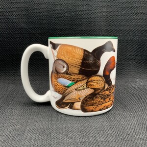 May include: White ceramic mug featuring a detailed illustration of various duck species. The mug has a green rim and a white handle. The ducks are depicted in shades of brown, black, orange, and blue. A perfect gift for bird watchers.