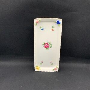 May include: A rectangular white ceramic tray with a scalloped edge. The tray is decorated with a pink rose in the center and small floral accents in pink, blue, yellow, and orange. The tray is approximately 8 inches long.