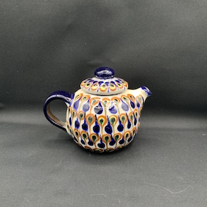 May include: A ceramic teapot featuring a cream-colored base decorated with a repeating pattern of blue, orange, and green teardrop shapes. The teapot includes a matching lid with a dark blue knob, a curved handle, and a short spout, all finished in a glossy glaze.