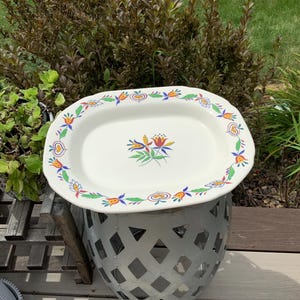 Könnte beinhalten: Eine cremeweiße, rechteckige Servierplatte mit gewelltem Rand, verziert mit einem bunten Blumen- und Herzmuster entlang des Randes sowie einem zentralen Motiv aus stilisierten Blumen und grünen Blättern in Rot, Gelb, Blau und Grün.