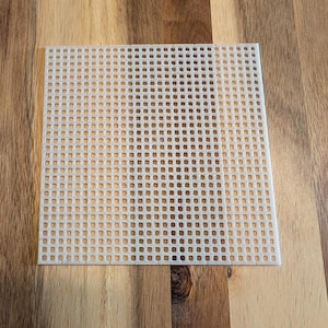 Puede incluir: Una lámina cuadrada de plástico de color blanco roto con agujeros cuadrados espaciados uniformemente. La lámina está colocada sobre una superficie de madera, mostrando su estructura de rejilla, comúnmente utilizada para proyectos de punto de aguja y punto de cruz.