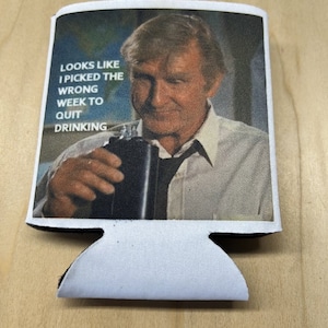 May include: White can cooler with a black trim and a photo of a man holding a glass of liquid. The text on the cooler reads "Looks like I picked the wrong week to quit drinking."