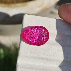 Puede incluir: Una gema facetada en forma de l&aacute;grima, de un vibrante color magenta. La gema presenta intrincados cortes radiales que emanan de un punto central, creando un efecto de estallido estelar. La piedra se muestra sobre una superficie blanca.