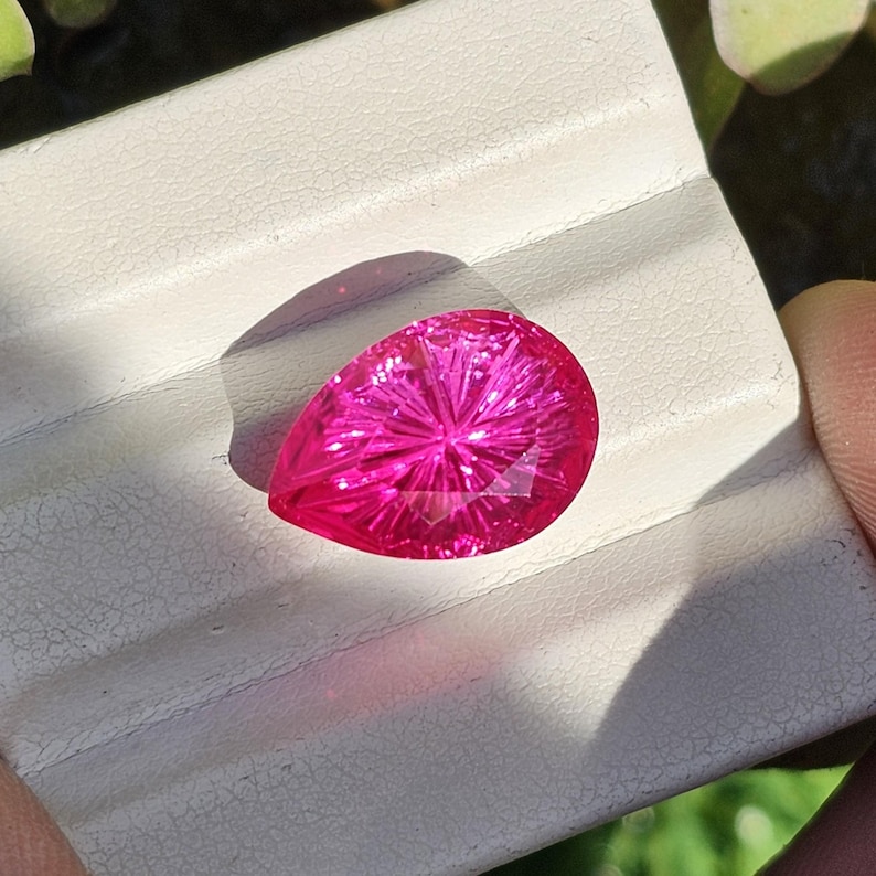 Puede incluir: Una gema facetada en forma de pera, de color magenta vibrante. El interior de la gema presenta un patr&oacute;n de estrella, que captura la luz. La piedra se sostiene sobre una superficie blanca, con una sombra a la izquierda.