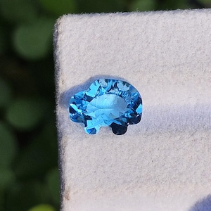 May include: A faceted, light blue gemstone shaped like a car. The gemstone has a bright, reflective surface and is set against a white background. The car shape has two small, dark wheels.