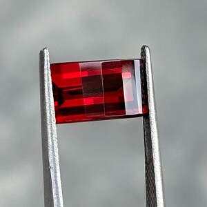 May include: A rectangular, faceted, deep red gemstone is held by silver tweezers. The gem's cut reveals internal reflections, enhancing its vibrant colour. The background is a soft, neutral grey.