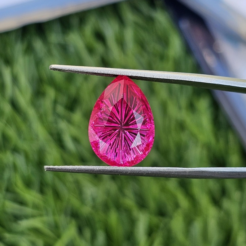 Puede incluir: Una gema facetada en forma de l&aacute;grima, de color rosa vibrante. La gema es sostenida por unas pinzas sobre un fondo verde borroso. El corte de la piedra crea un patr&oacute;n de estrella dentro de la gema.