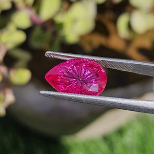 Puede incluir: Una gema facetada en forma de pera, de color magenta vibrante, es sostenida por unas pinzas. Los intrincados cortes de la gema crean un patr&oacute;n de estrella, reflejando la luz. El fondo es un suave desenfoque de follaje verde.