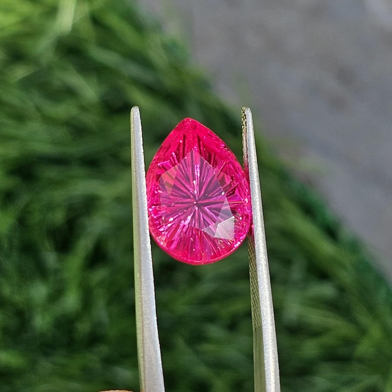 Puede incluir: Una gema rosa intenso facetada en forma de l&aacute;grima es sostenida por unas pinzas plateadas sobre un fondo verde borroso. La gema tiene un patr&oacute;n de cortes en forma de estrella que irradia desde el centro.