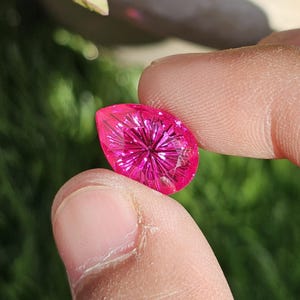 Puede incluir: Una gema facetada en forma de l&aacute;grima, de un vibrante color magenta. La gema se sostiene entre dos dedos, mostrando sus intrincados cortes internos que crean un efecto de estallido estelar. El fondo est&aacute; borroso, con follaje verde.