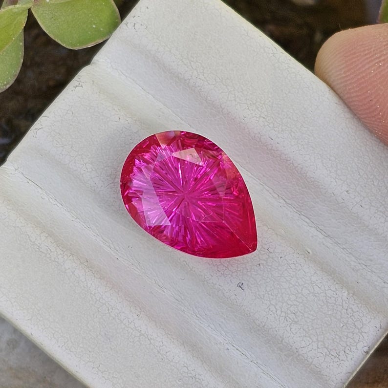Puede incluir: Una gema facetada en forma de l&aacute;grima, de un vibrante color magenta. El interior de la gema presenta un patr&oacute;n radial, creando un efecto de estallido estelar. La piedra se encuentra sobre una superficie blanca, con un toque de vegetaci&oacute;n en el fondo.
