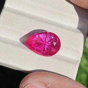 Puede incluir: Una gema facetada en forma de pera, de un vibrante color magenta. Los intrincados cortes crean un patr&oacute;n de estrella, capturando la luz. La piedra se muestra sobre una superficie blanca, con un toque de hierba verde al fondo.