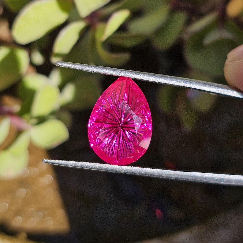 Puede incluir: Una gema facetada en forma de l&aacute;grima, de color rosa intenso, es sostenida por unas pinzas. La gema tiene un patr&oacute;n de estrella que irradia desde el centro. Se ve follaje verde en el fondo.