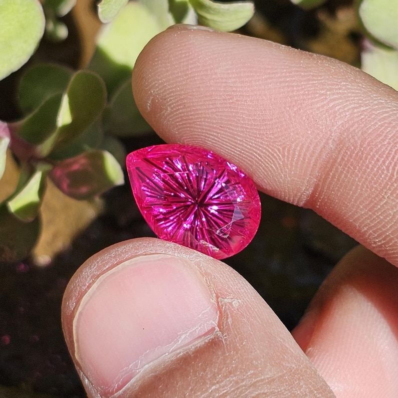 Puede incluir: Una gema facetada en forma de l&aacute;grima, de un vibrante color magenta. La gema se sostiene entre dos dedos, mostrando su intrincado dise&ntilde;o interno. El corte crea un patr&oacute;n de estrella que irradia desde el centro.