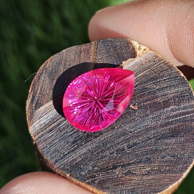 Puede incluir: Una gema facetada en forma de l&aacute;grima, de un vibrante color magenta. La gema est&aacute; colocada sobre un trozo de madera oscura y texturizada. El corte de la piedra crea un patr&oacute;n de estrella. El fondo es verde borroso.