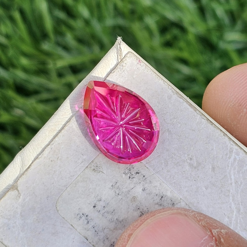 Puede incluir: Una gema facetada en forma de l&aacute;grima de color rosa vibrante. La gema presenta intrincadas tallas en forma de estrella en su superficie. La piedra se sostiene contra una superficie blanca, con un fondo verde visible.