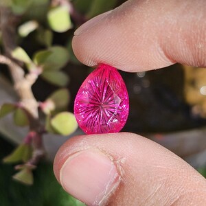 Puede incluir: Una gema facetada en forma de l&aacute;grima, de color magenta vibrante. La gema se sostiene entre dos dedos, mostrando su intrincado patr&oacute;n interno de estrella. El fondo est&aacute; borroso, con indicios de follaje verde.