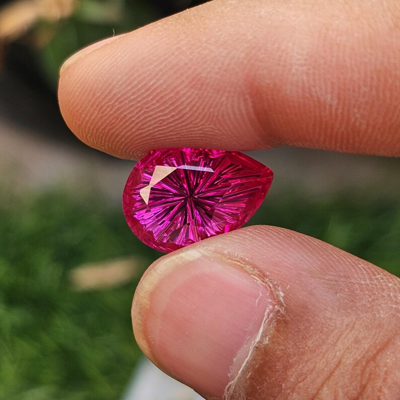 Puede incluir: Una gema facetada en forma de l&aacute;grima, de un vibrante color magenta. La gema se sostiene entre dos dedos, mostrando su intrincado patr&oacute;n interno de l&iacute;neas radiales. El corte de la piedra crea un efecto de estallido estelar.