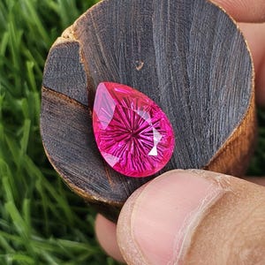 May include: A faceted, teardrop-shaped gemstone in a vibrant magenta hue. The gem features intricate internal cuts radiating from a central point, creating a starburst effect. The gem is set against a dark, textured wooden surface.