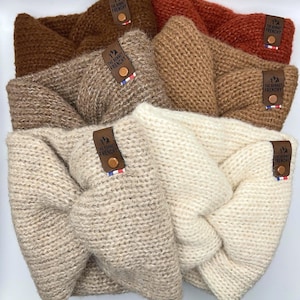 Puede incluir: Una colección de diademas de punto en varios colores neutros, crema, beige y marrón. Cada diadema presenta un diseño retorcido y una pequeña etiqueta de cuero con el texto "THE BONNET FRENCHY". Las diademas están hechas de un hilo grueso y texturizado.