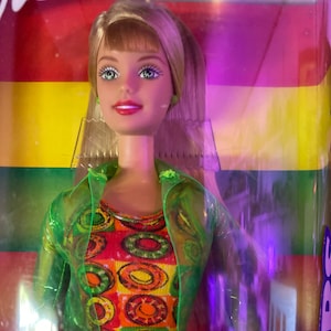 May include: A Barbie doll with blonde hair and green eyes wearing a green jacket and a colorful dress with a circle pattern. The doll is packaged in a box with a rainbow background.